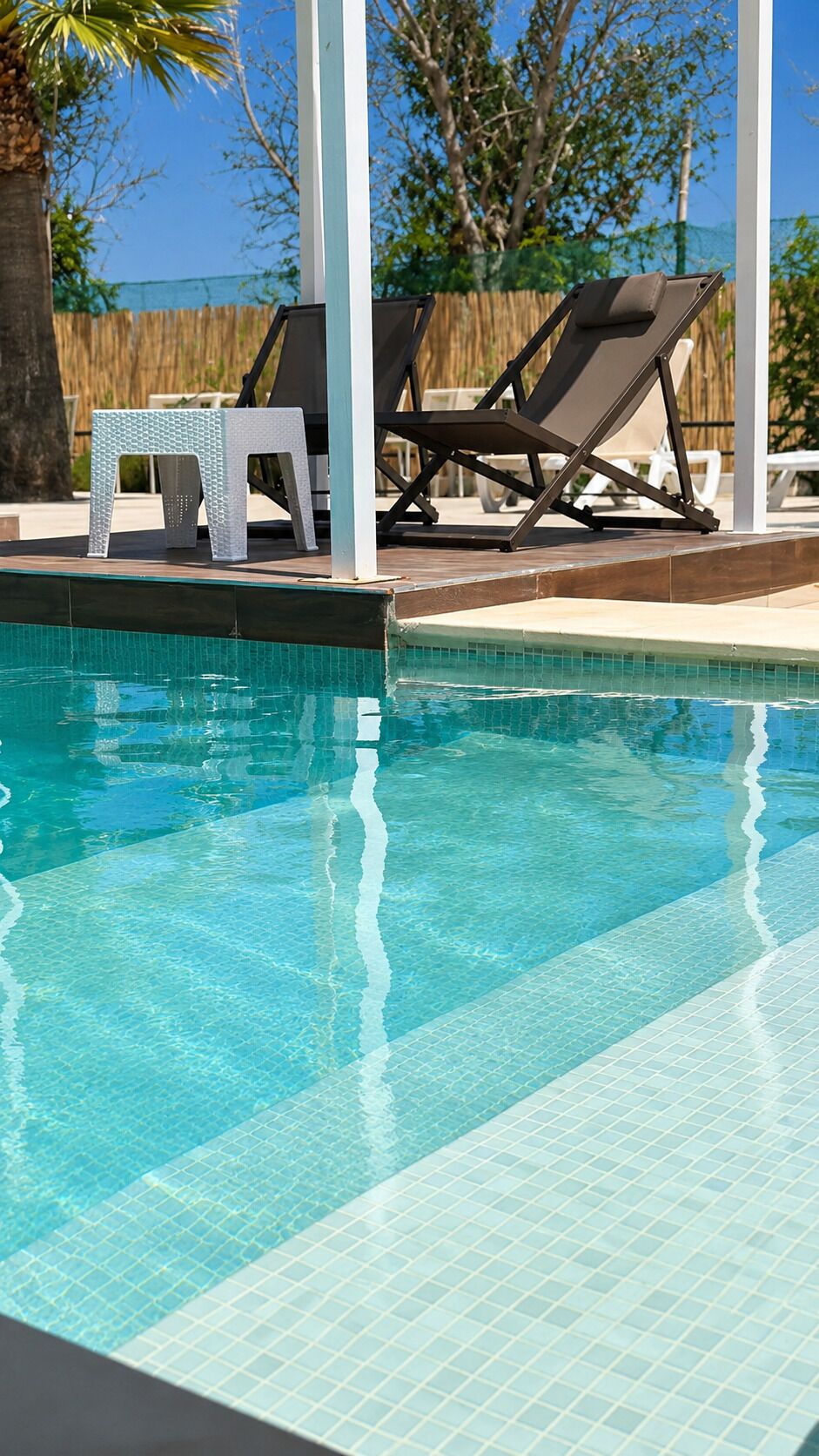 A clear swimming pool with lounge chairs and a small table, surrounded by a wooden deck and a fence.