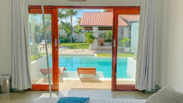 A room with a large sliding glass door leading to a pool area, a bed with a blue throw, and a wicker chair by the pool. - Kite Soul Pousada (Caucaia)