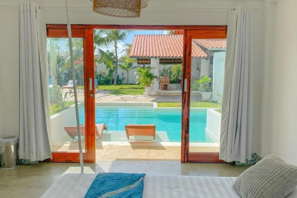 Quarto com uma grande porta de vidro deslizante que dá para uma área de piscina, uma cama com manta azul e uma cadeira de vime junto à piscina.