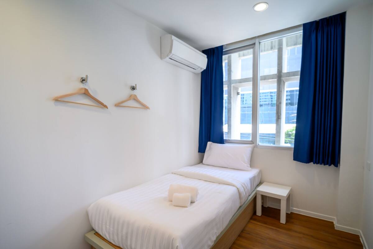 A bedroom with a bed, a bedside table, a hanging air conditioner, and a window with curtains.
