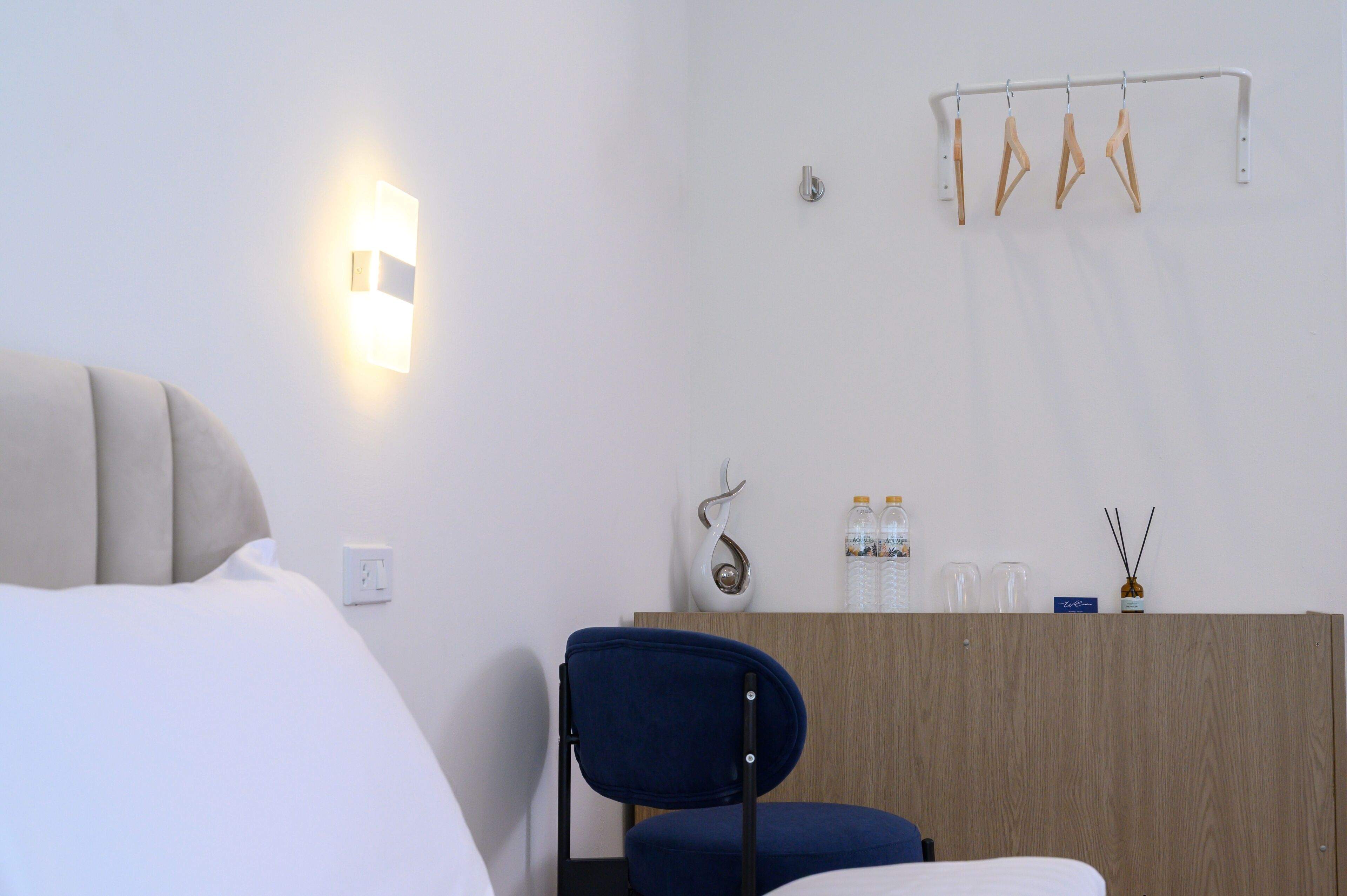 A hotel room with a bed, a desk with a chair, a wall-mounted light, and a coat rack.