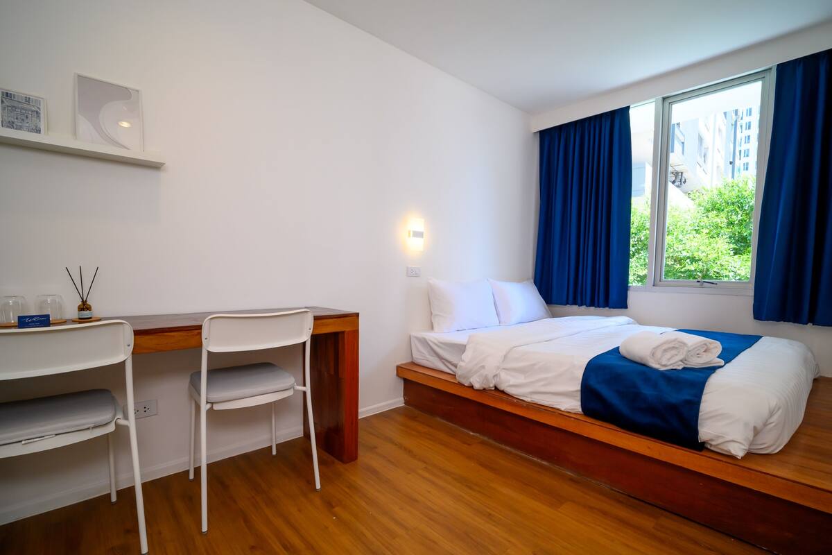 A bedroom with a wooden bed, white bedding, a desk with a chair, and a window with blue curtains.