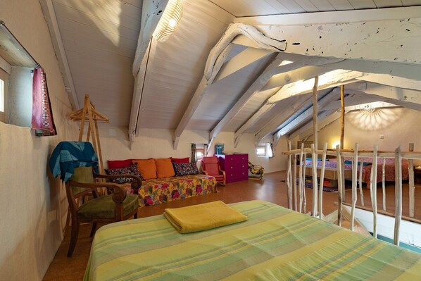 Una habitación con techo inclinado, una cama con edredón a rayas, una silla de madera, un sofá con cojines de colores y una mesita.