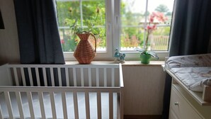 Una cuna con barandilla blanca, un aparador con cajones, una ventana con vistas al verdor, una cesta de mimbre y una pequeña figurita en el alféizar.