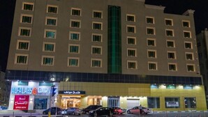 A multi-story hotel with a central green vertical feature, multiple ground-level shops, and a parking area with cars.
