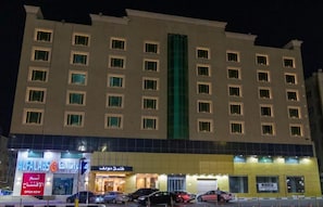 Un hotel de varios pisos con un elemento vertical verde en el centro, varias tiendas a nivel de calle y un estacionamiento con automóviles.