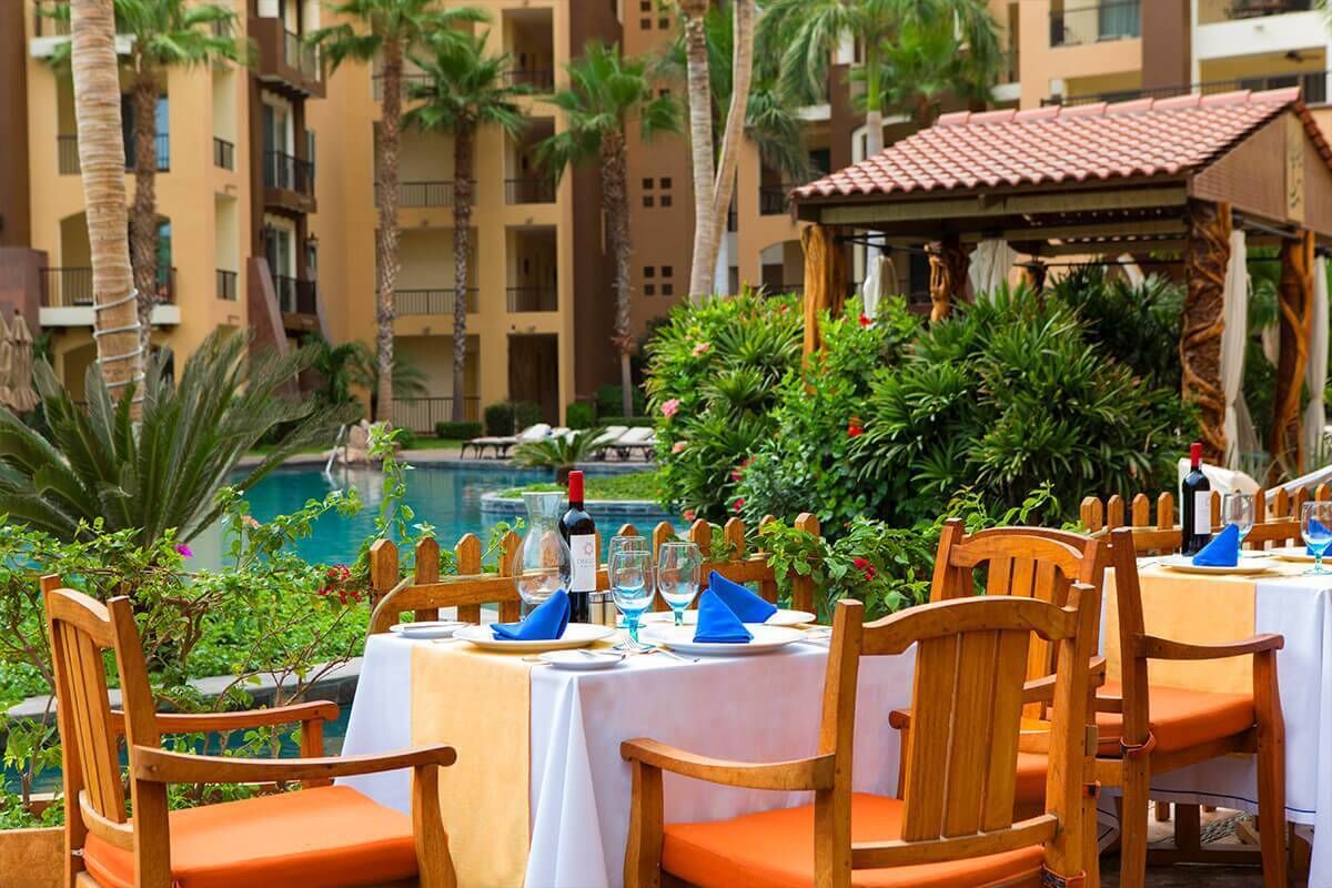 A dining area with tables set for four, surrounded by lush greenery and a pool in the background.