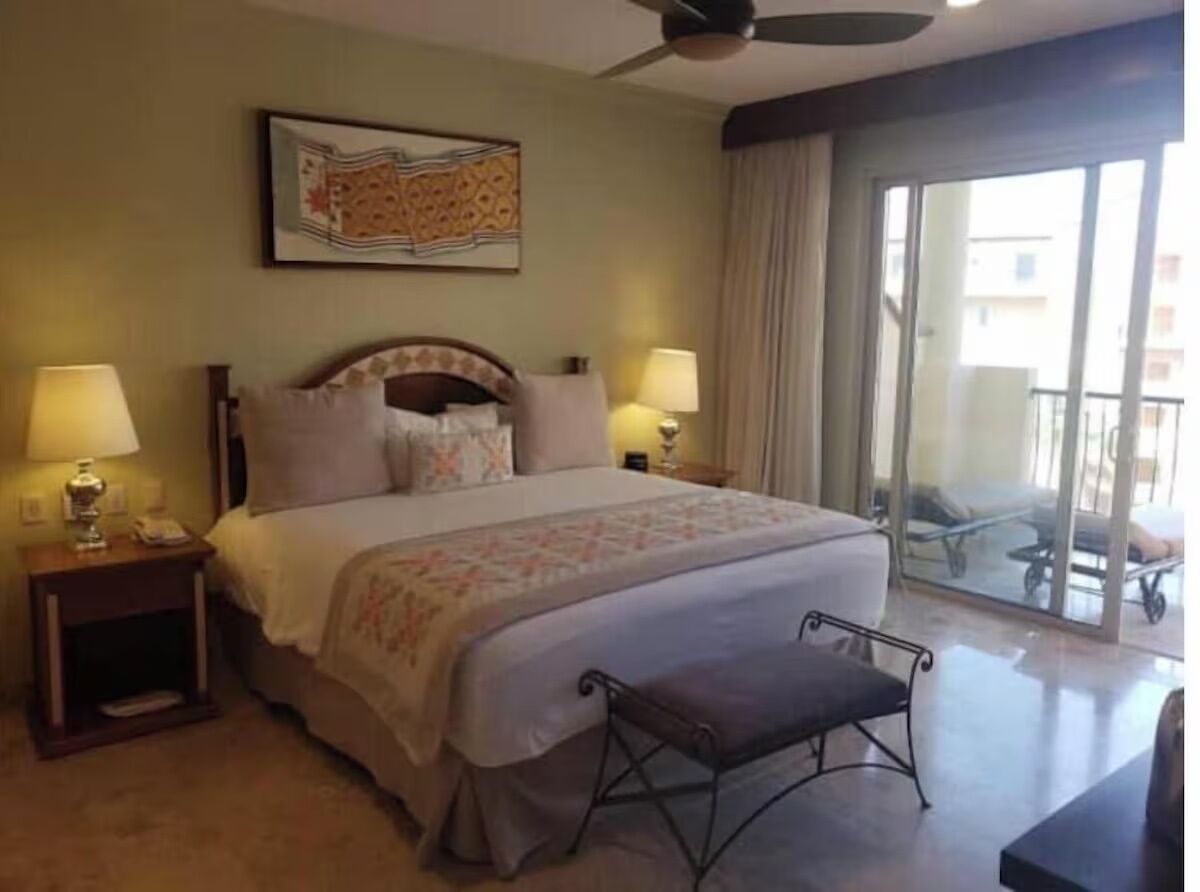A hotel room with a bed, two bedside lamps, a ceiling fan, a large window with a view, and a small bench.