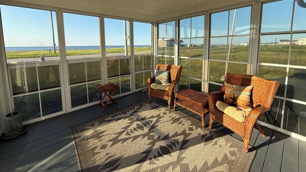 A covered porch with wicker furniture, a patterned rug, and large windows offering a view of the ocean and farmland.