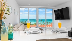 A dining area with a table set for two, a TV mounted on the wall, and a view of the ocean through large windows.