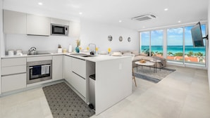 A modern kitchen with a central island, a built-in oven, and a microwave. There is a dining area with a sofa and a table, and large windows offering a view of the ocean.