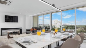 A modern dining area with a large table set for six, a sofa, a TV, and a city view through large windows.