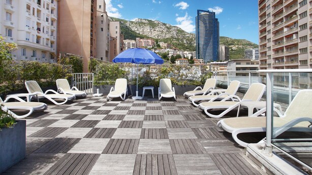 Une terrasse sur le toit, dotée de chaises longues et d’un parasol bleu, offrant une vue sur un paysage urbain avec ses bâtiments et une montagne.