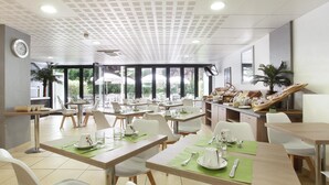 Un espace de restauration moderne, avec des tables dressées pour les repas, de grandes fenêtres offrant une vue sur la verdure, et un coin cuisine visible au fond.