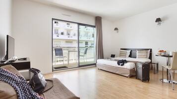 A modern hotel room with a bed, a desk, a chair, a TV, and a view of the city through large windows.