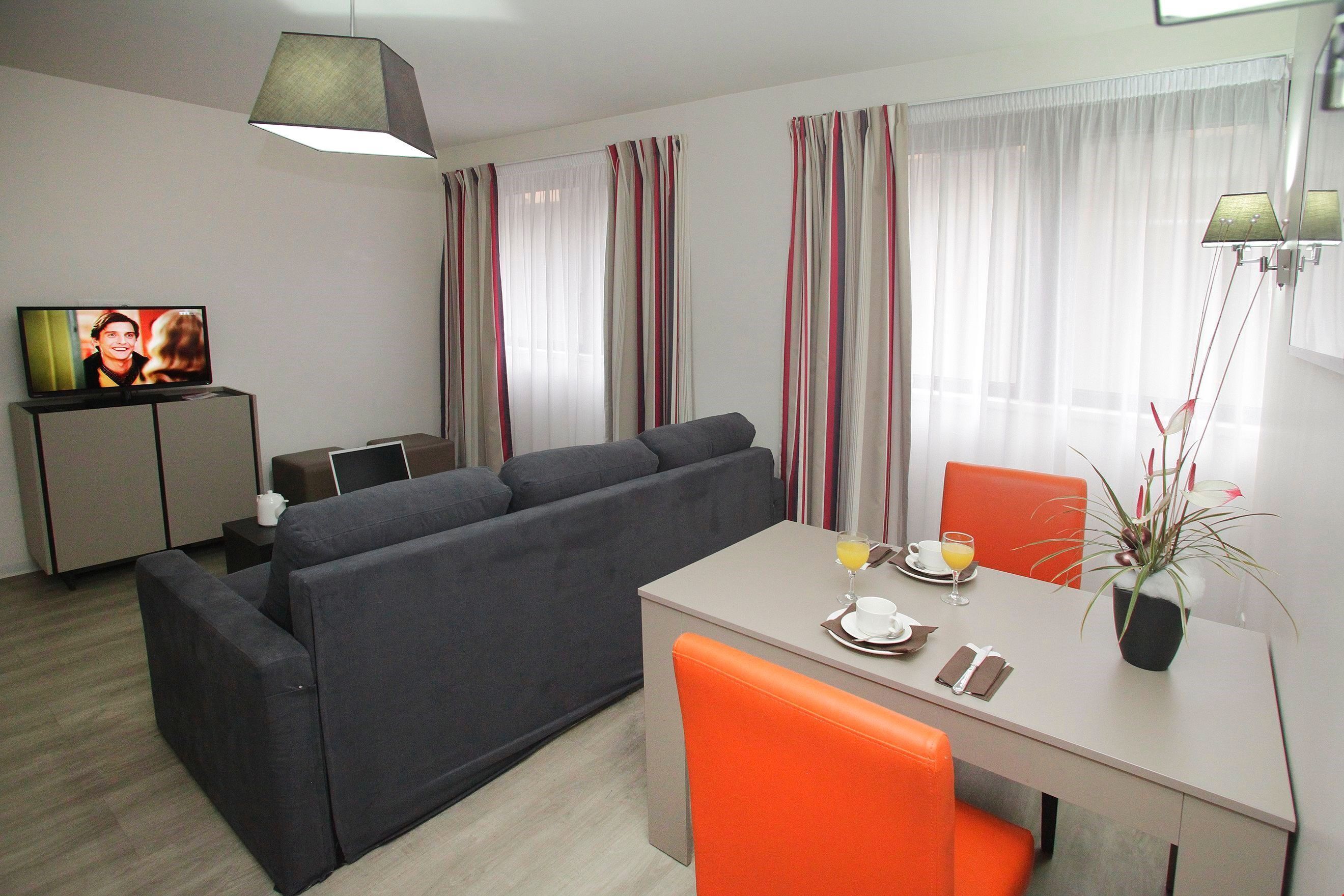 Un soggiorno moderno con un divano grigio, un tavolo da pranzo con sedie arancioni, una TV su un mobile e una pianta in vaso.