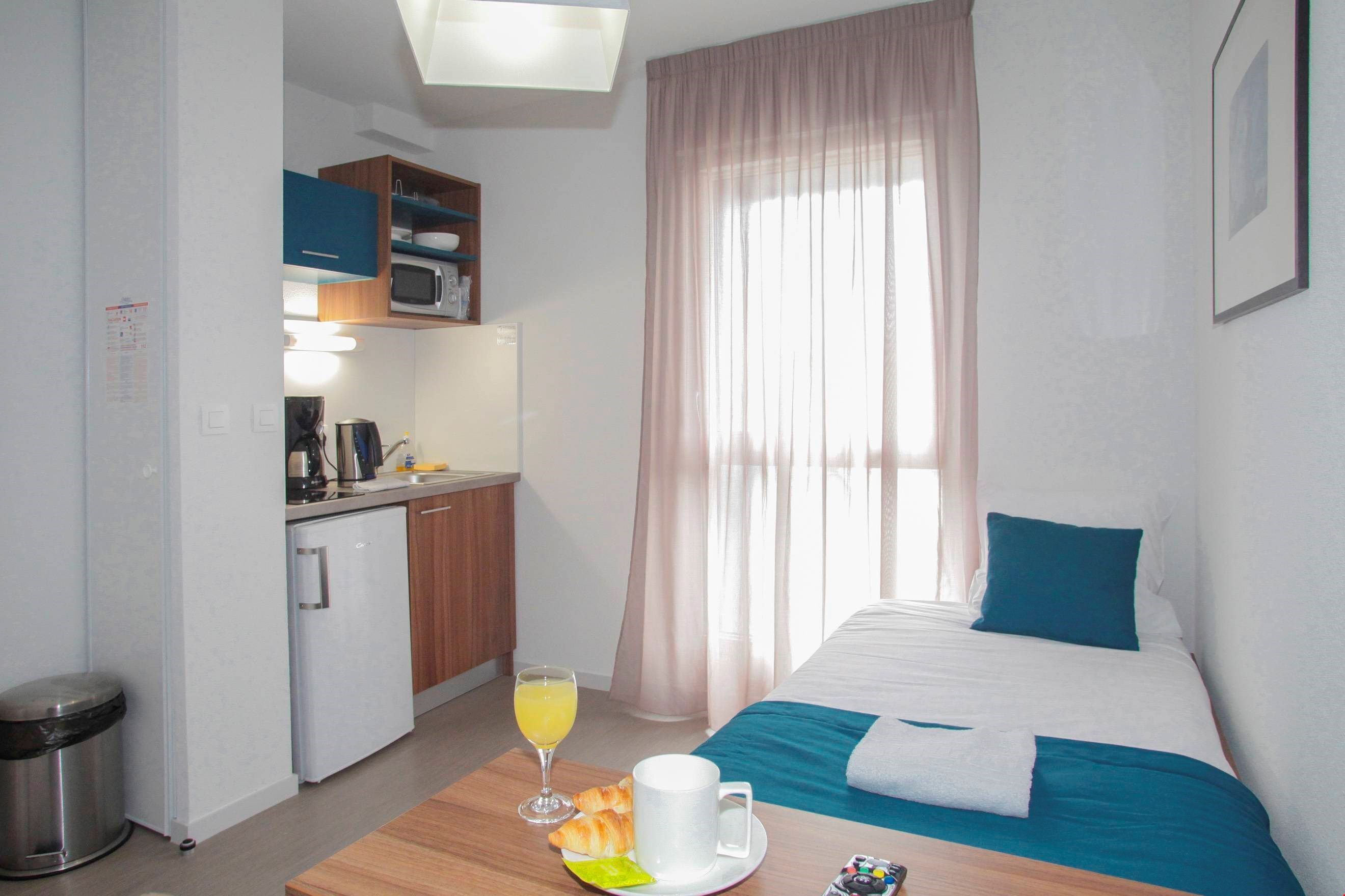 Hotelkamer met een bed, een kleine kitchenette, een eettafel met ontbijtartikelen en een raam met gordijnen.