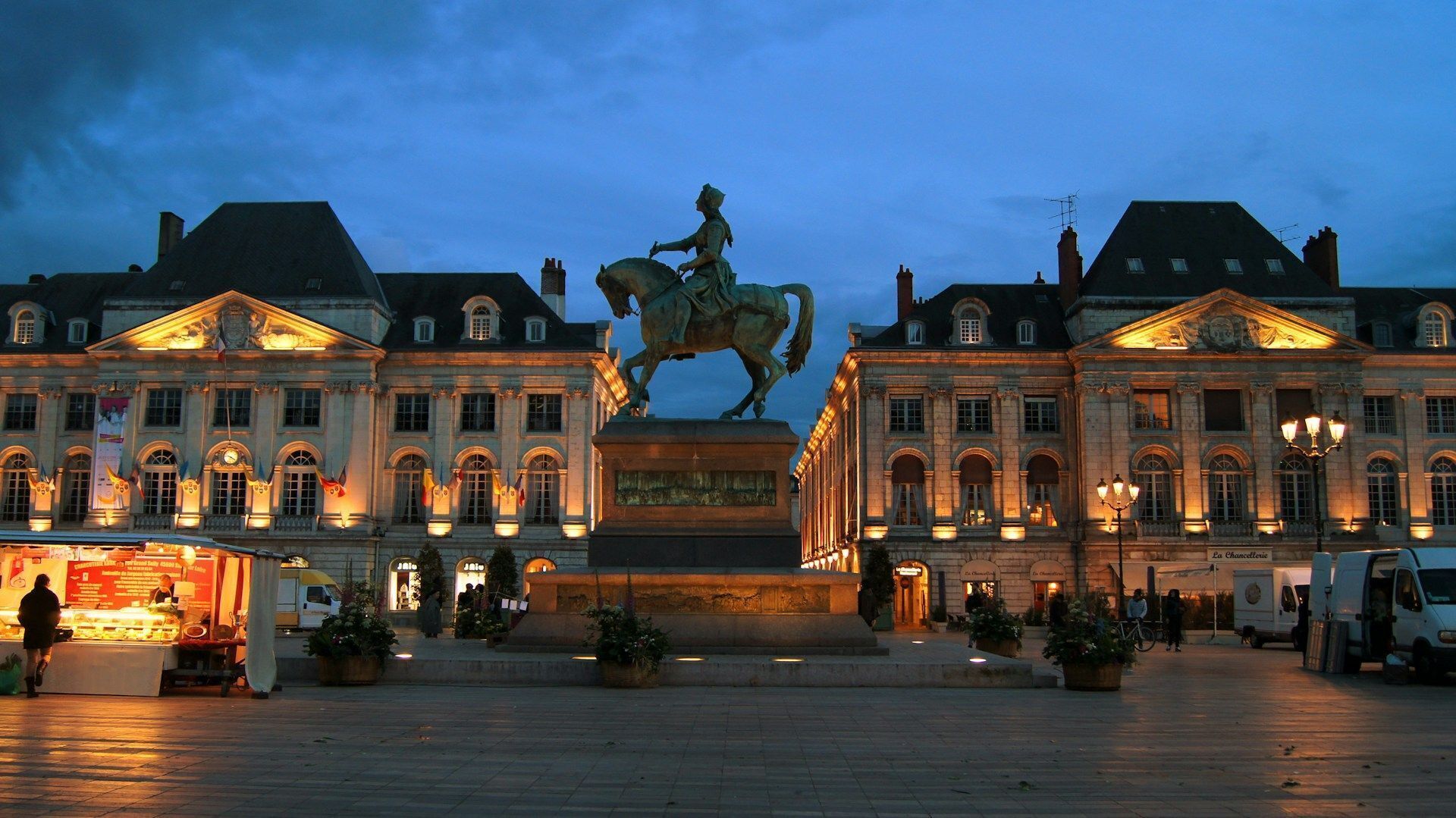 Un bâtiment historique doté d’une statue centrale, de grandes fenêtres et d’une place dallée.