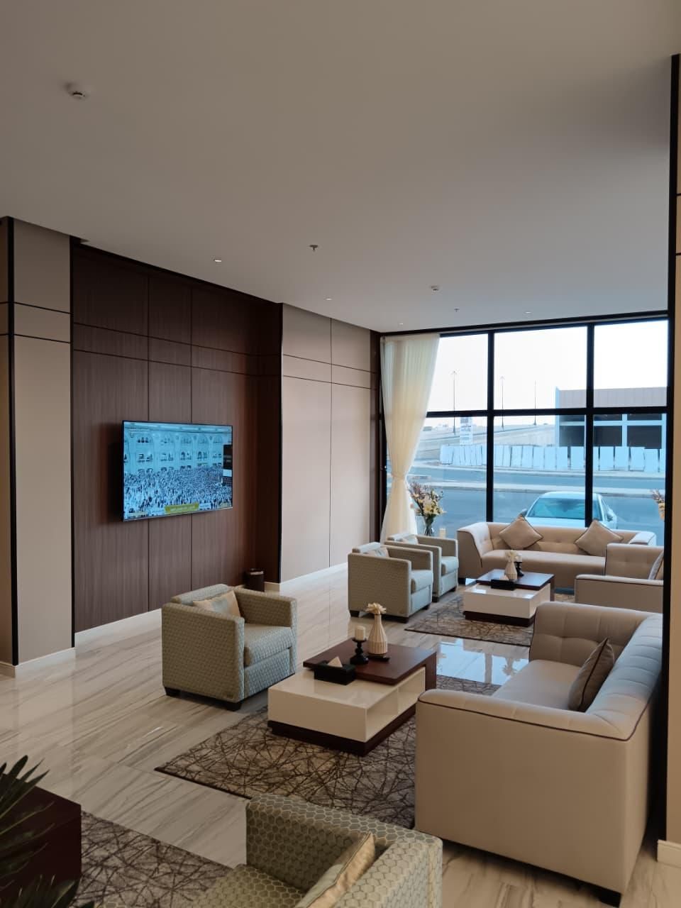 Eine moderne Hotelhalle mit einem großen Flachbildfernseher an der Wand, einer Sitzecke mit Sofas und Sesseln und einem großen Fenster mit Blick auf das Flughafengelände.