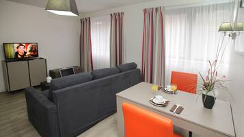 Ein modernes Wohnzimmer mit einem grauen Sofa, einem Esstisch mit orangefarbenen Stühlen, einem Fernseher auf einer Kommode und einem Topf mit Pflanze.