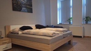 Ein Schlafzimmer mit Bett, Nachttischen, einem Heizkörper und Fenstern mit Pflanzen.