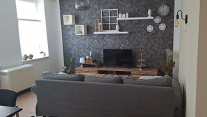 Ein Wohnzimmer mit grauem Sofa, einem Fernseher auf einem Holzständer, dekorativen Wandschränken und einem Fenster mit Jalousien.