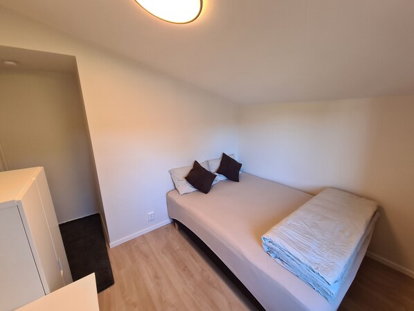 Une petite chambre bien éclairée, avec un lit simple, une table de chevet et un luminaire fixé au mur.