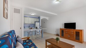A living room with a sofa, a coffee table, a TV on a wooden cabinet, and a kitchen area with a fridge and bar stools. - Brisas de Chasna Holiday Apartment with Sea Views, Shared Pool and Wi-Fi (Costa Adeje)