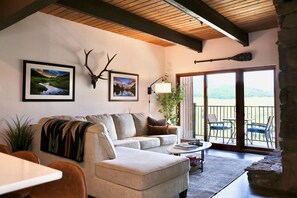 A cozy living room with a sofa, a coffee table, and a dining table. There are framed pictures on the wall, a plant, and a stone fireplace.