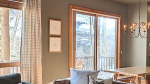 Un salon chaleureux avec un canapé en cuir marron, un fauteuil vert et une table basse en bois. Il y a deux fenêtres avec des rideaux, un lustre et des tableaux encadrés accrochés au mur.