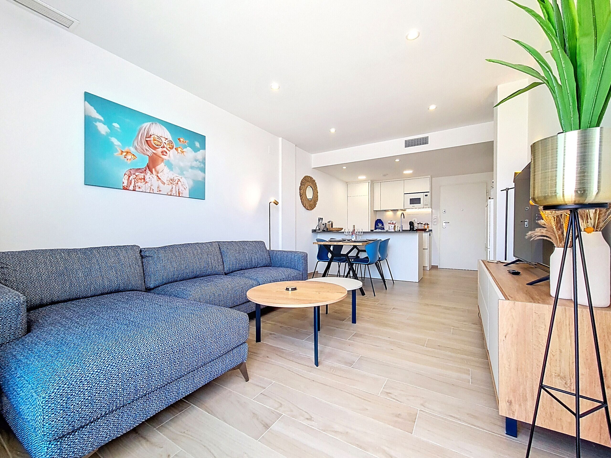 A modern living room with a blue sofa, a round coffee table, and a dining area with blue chairs. There is a kitchen in the background with white cabinets and a microwave.
