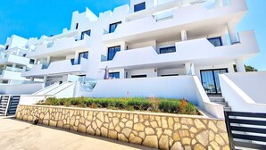A modern multi-story building with balconies, a stone retaining wall, and a clear blue sky.