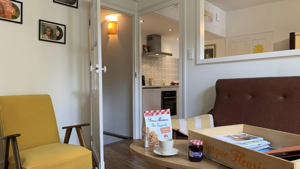 Una sala de estar con un sillón amarillo, un sofá marrón, una mesa de centro de madera y una cocina a la vista a través de una puerta abierta.