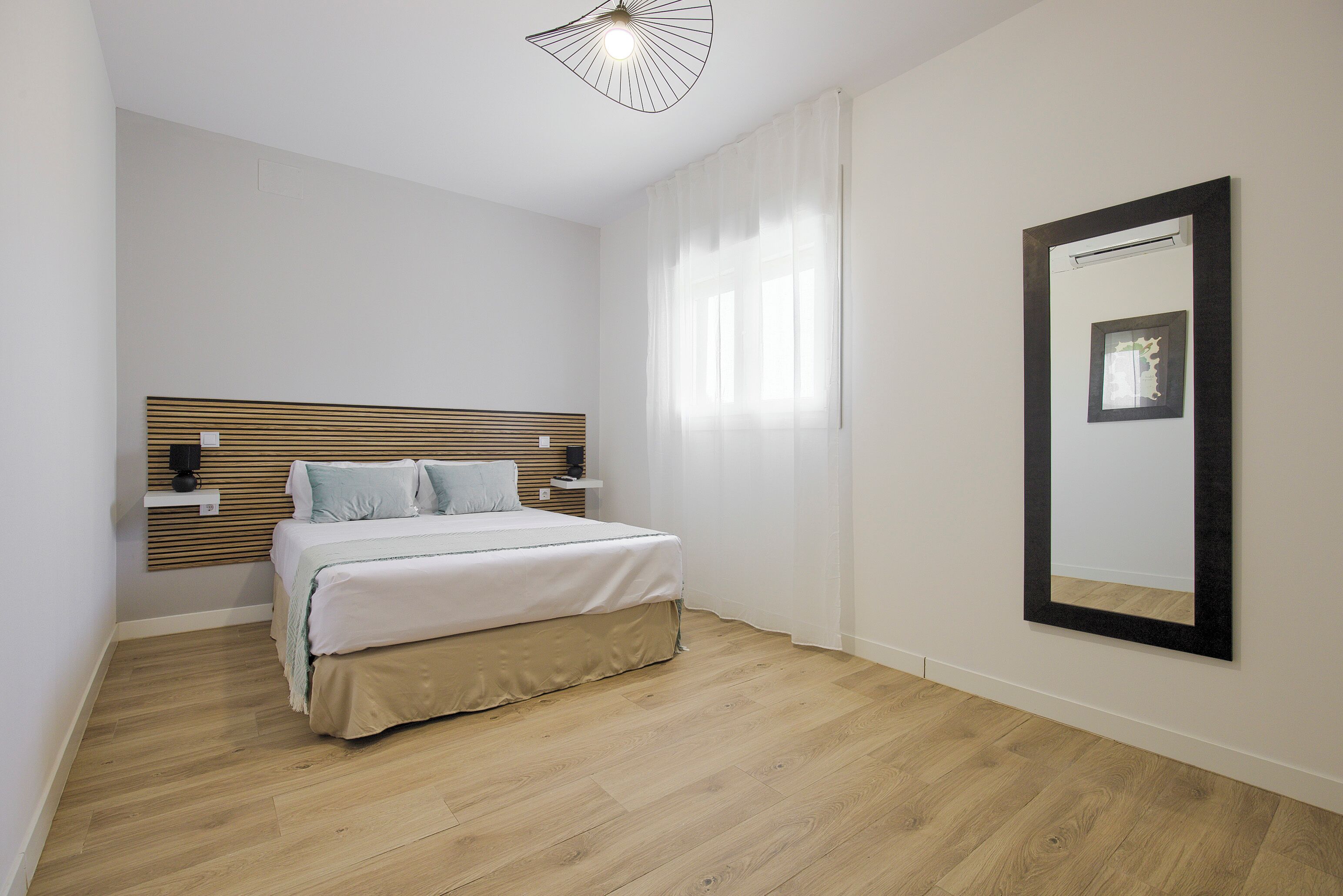 Un dormitorio con cabecera de madera, cama con ropa de cama blanca, mesita de noche y un espejo grande en la pared.