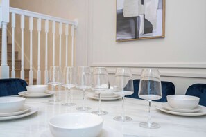 Mesa de jantar posta com pratos, tigelas e copos de vinho brancos, com uma escadaria e um quadro emoldurado ao fundo.