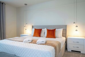 Cama bem arrumada com roupa de cama branca e laranja, duas mesas de cabeceira com abajures e cabeceira cinza.