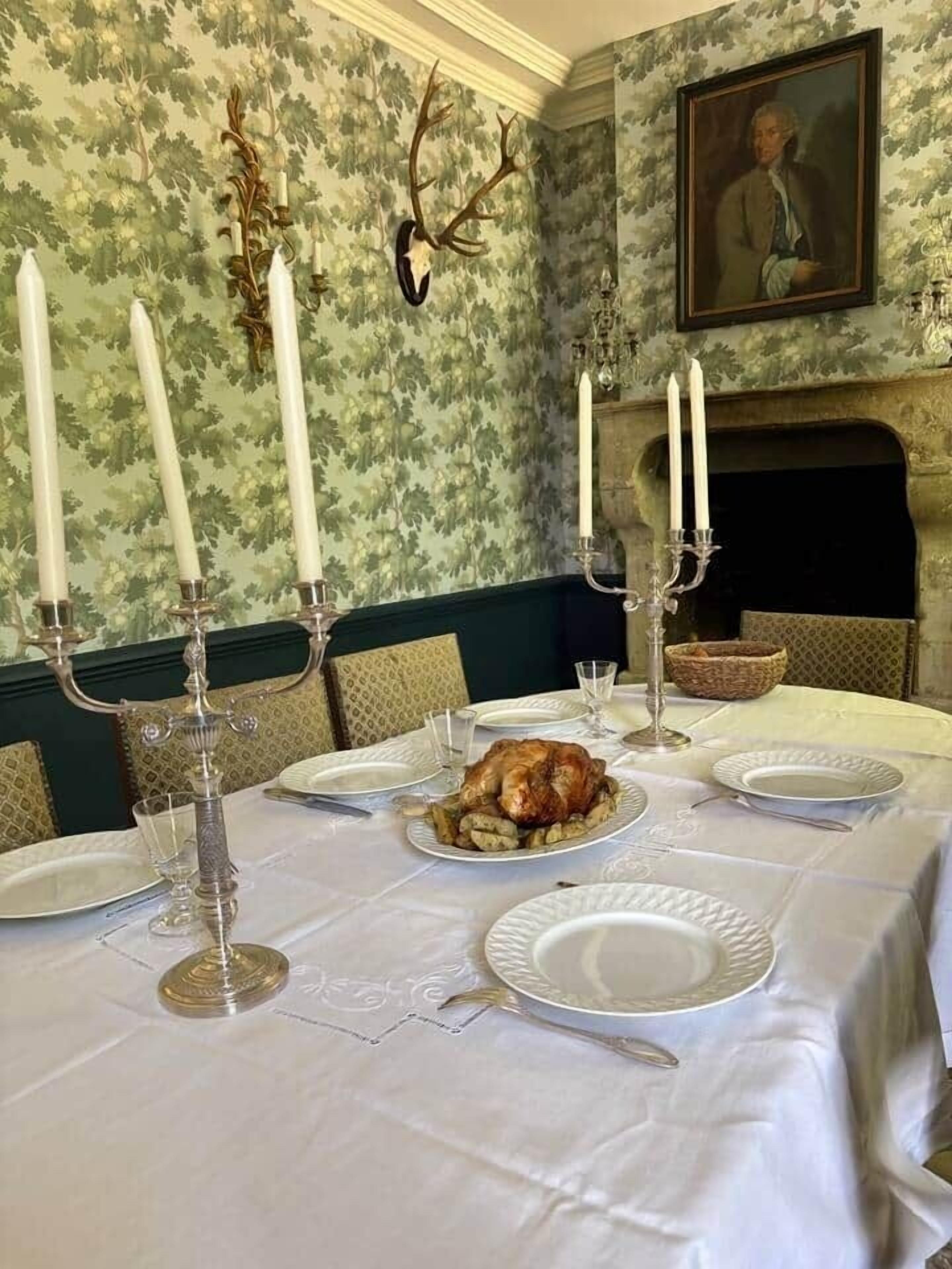 Une salle à manger avec une table dressée pour un repas, agrémentée d’un poulet rôti en pièce maîtresse, de vaisselle blanche et de chandeliers.