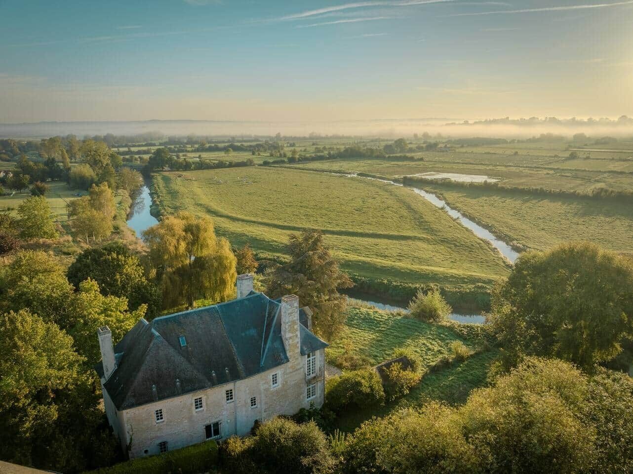 Een groot stenen gebouw met een steil dak, omgeven door weelderige begroeiing en een rivier.