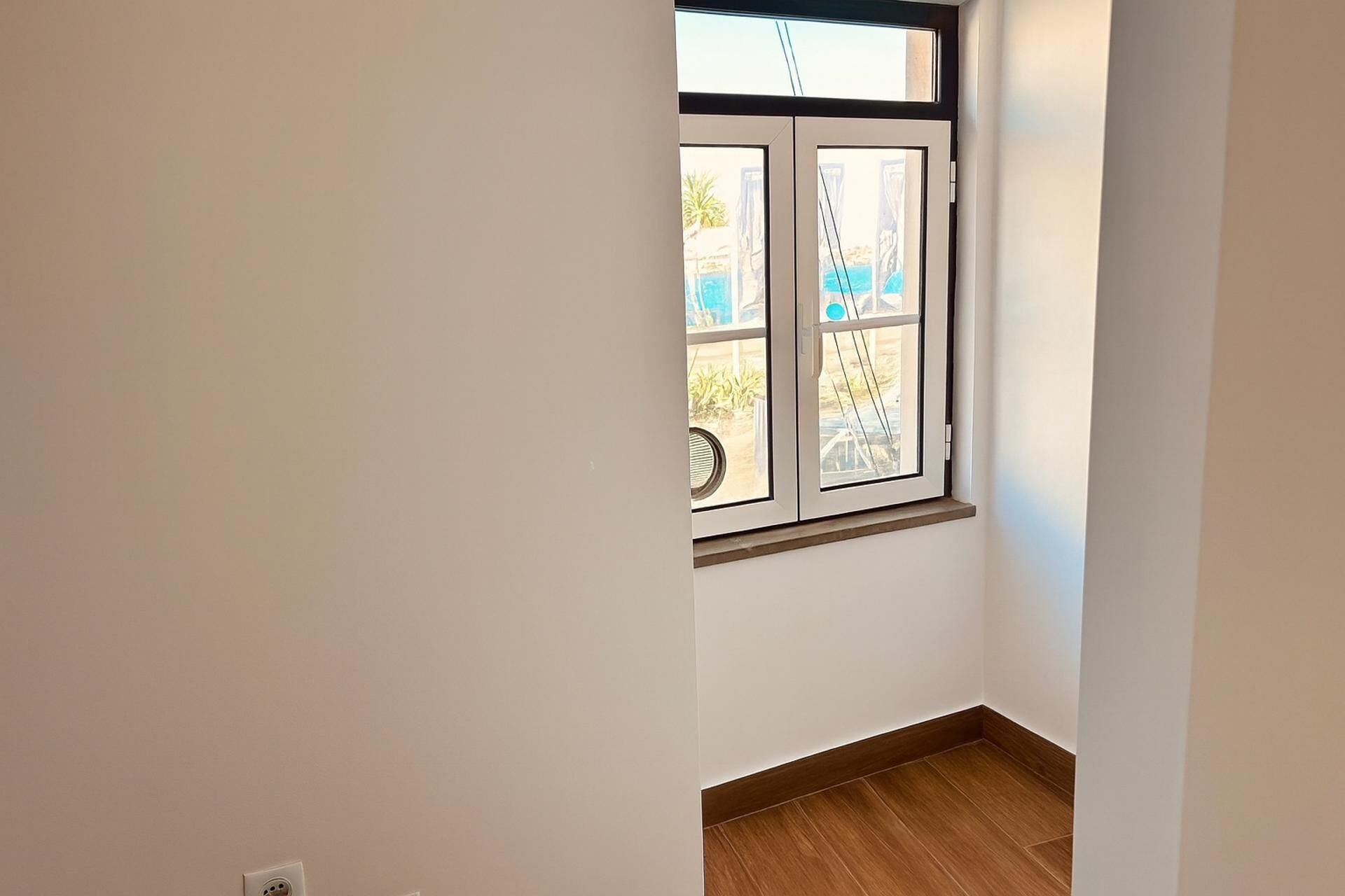 Una habitación con ventana, suelo de madera y pared blanca.