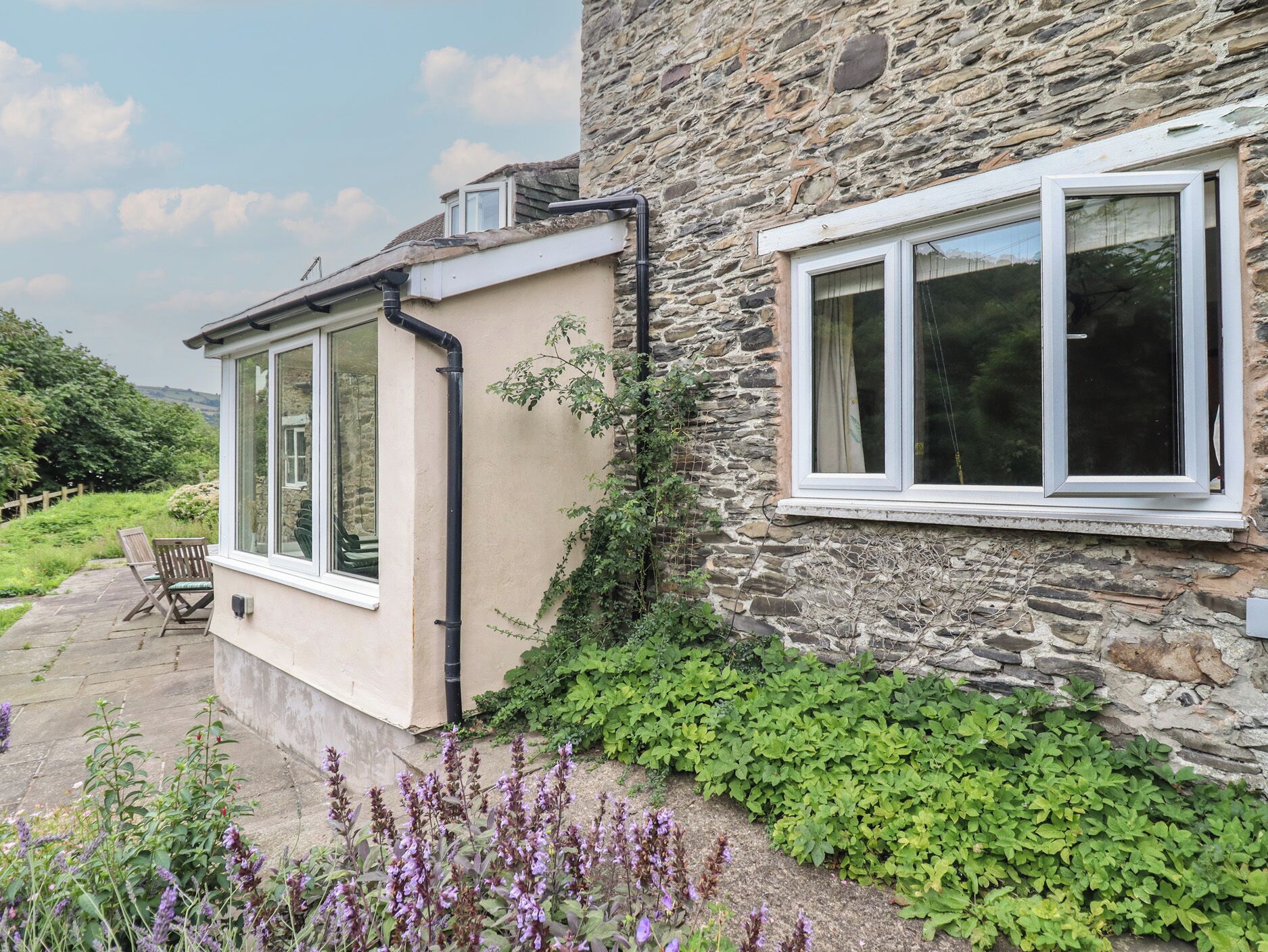 Ein Steinhaus mit Fenster und Tür, ein hölzerner Stuhl und lila Blumen im Vordergrund.