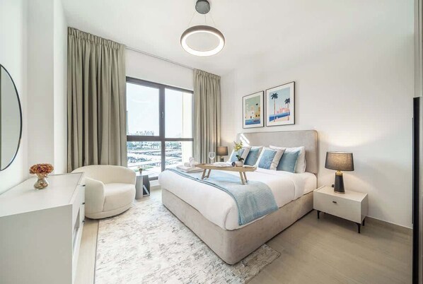 A modern hotel room with a large bed, a desk, a chair, a nightstand, and a window with curtains.