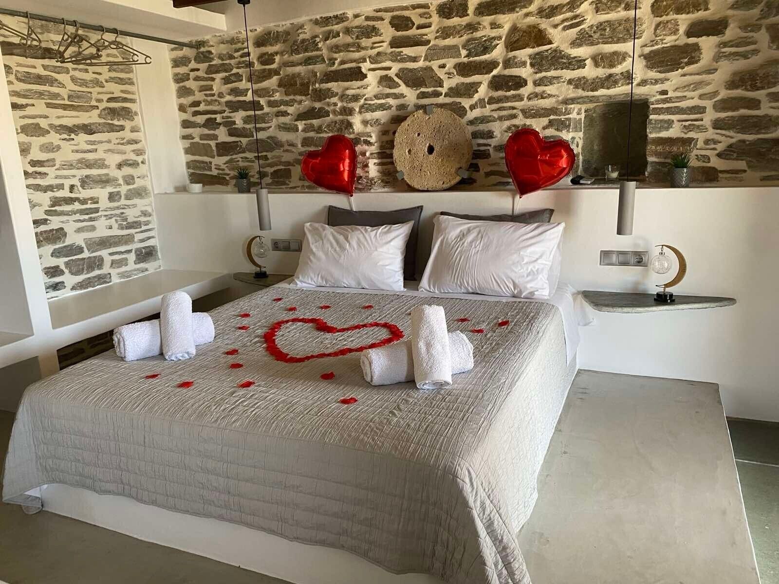 A bed with a heart-shaped towel arrangement, a stone wall, and a bedside table with a lamp.