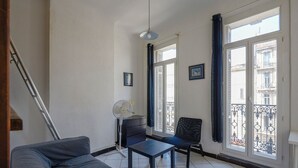 A living room with a sofa, a table, a chair, a fan, and a window with a view of buildings.