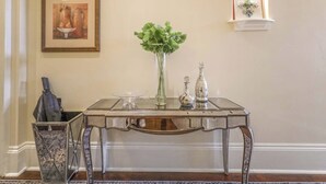A glass-top table with a vase of green foliage, two silver vases, and a framed artwork on the wall. - Charming Condo w Patio off Magazine St 2br|1ba (New Orleans)