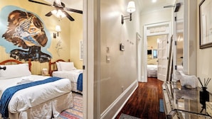 A hotel room with a bed, a ceiling fan, a wall-mounted lamp, a door, and a hallway leading to another room. - Charming Condo w Patio off Magazine St 2br|1ba (New Orleans)