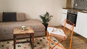 Een gezellige woonkamer met een bank, een klein houten tafeltje, een kamerplant en een keuken met een fornuis en kastjes.