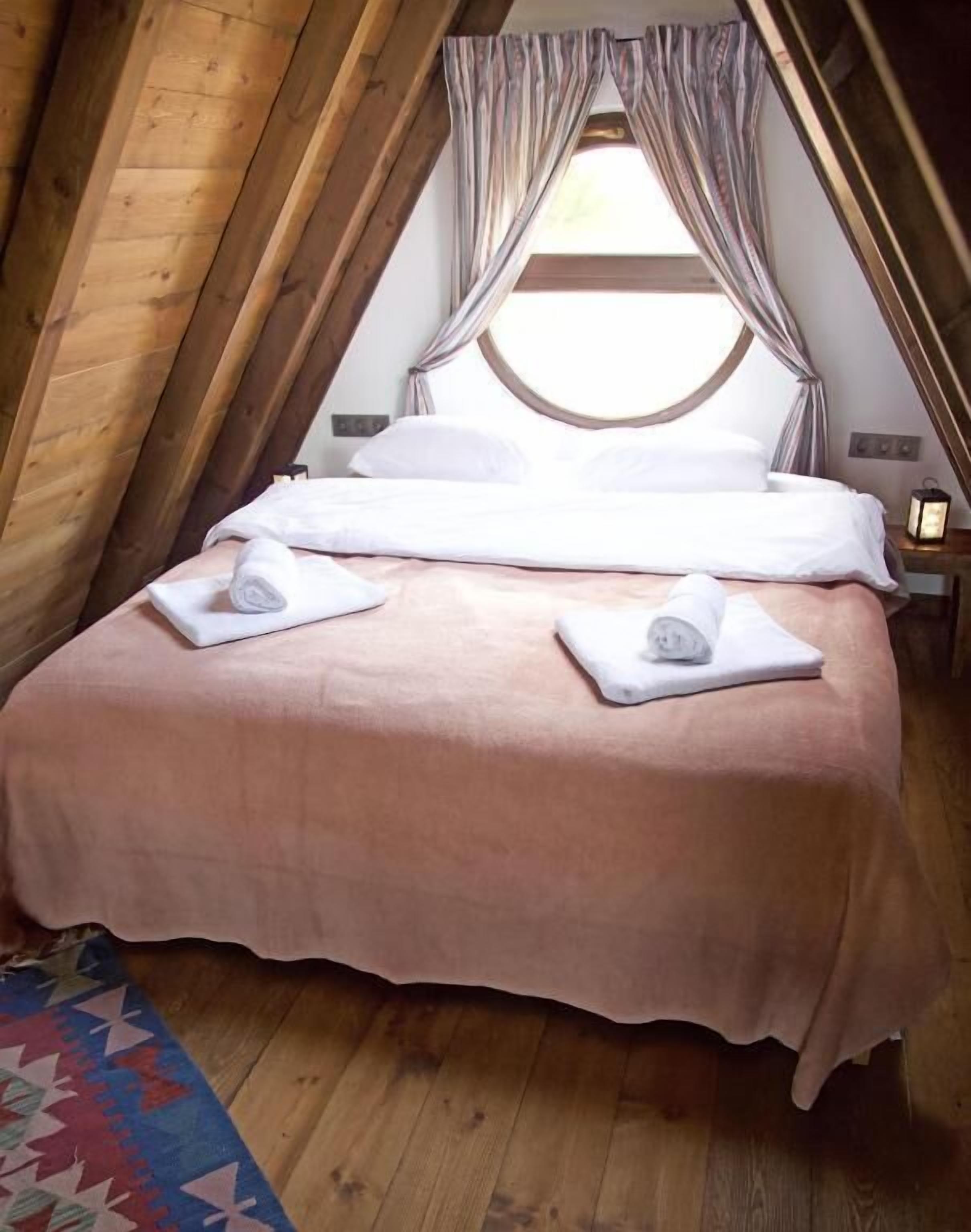 A cozy, single-bed room with a sloped ceiling, a large window with curtains, and a wooden floor.