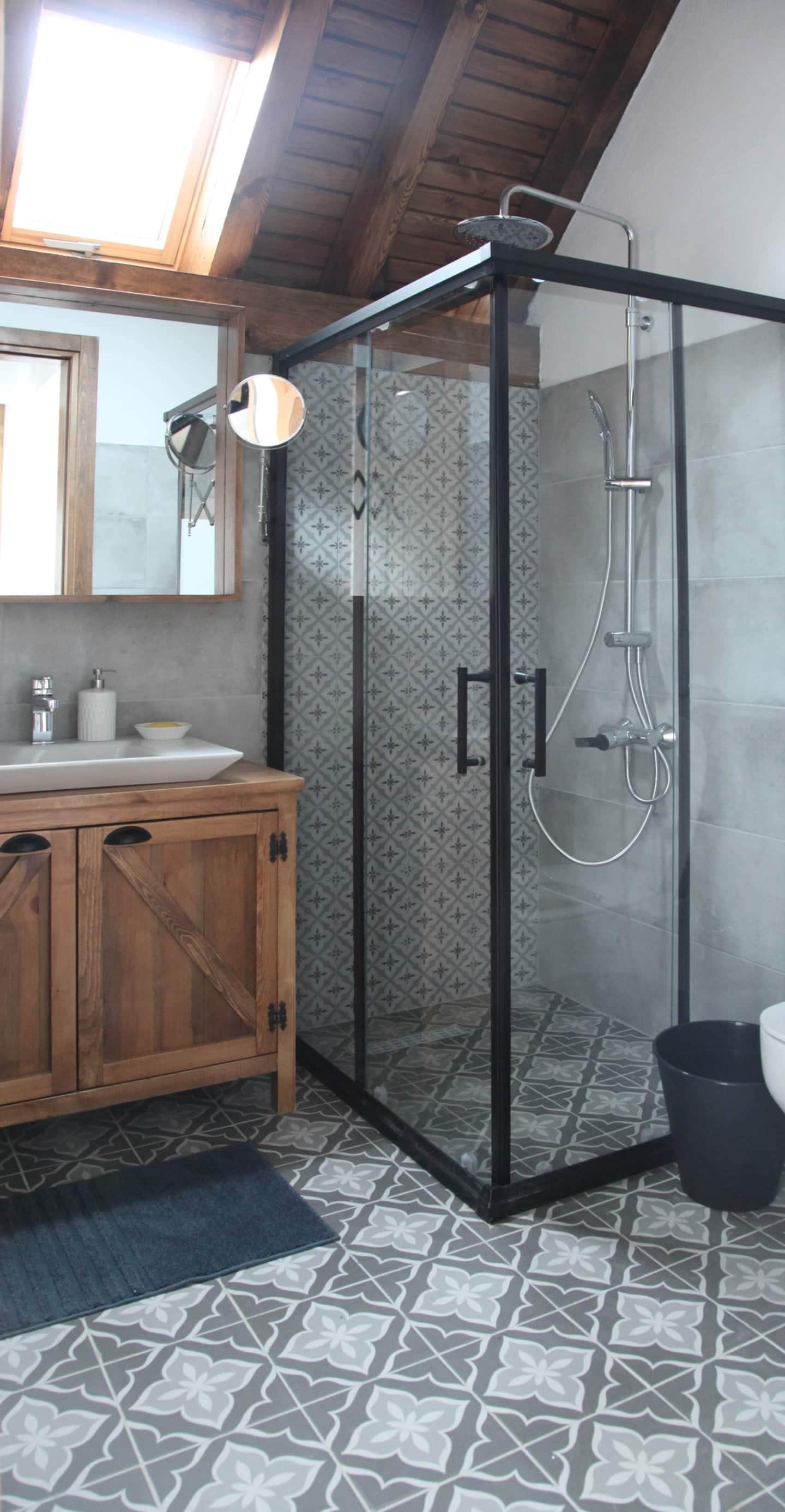 Una ducha con puerta de vidrio, un tocador de madera con espejo y baldosas de suelo con diseño.