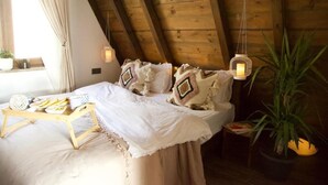 Een knus slaapkamer met een houten plafond, een bed met witte lakens, decoratieve kussens, een nachtkastje met een lamp, en een raam met gordijnen.