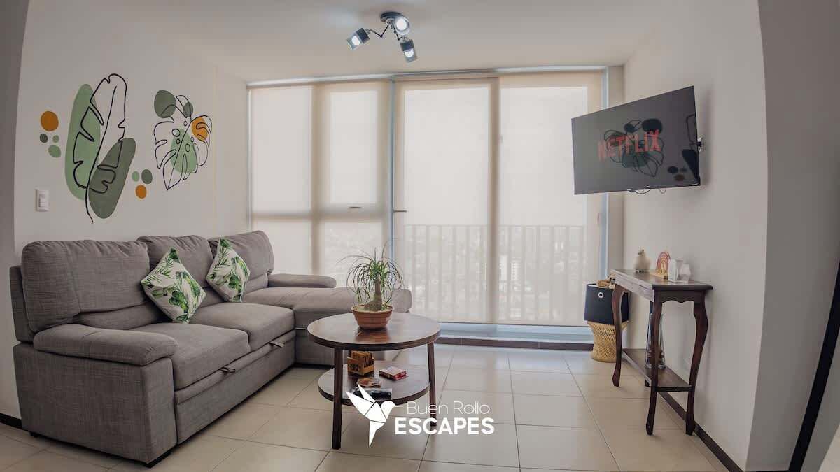 Una sala de estar con un sofá gris, una mesita redonda con una planta, un televisor montado en la pared y una puerta con salida al balcón.
